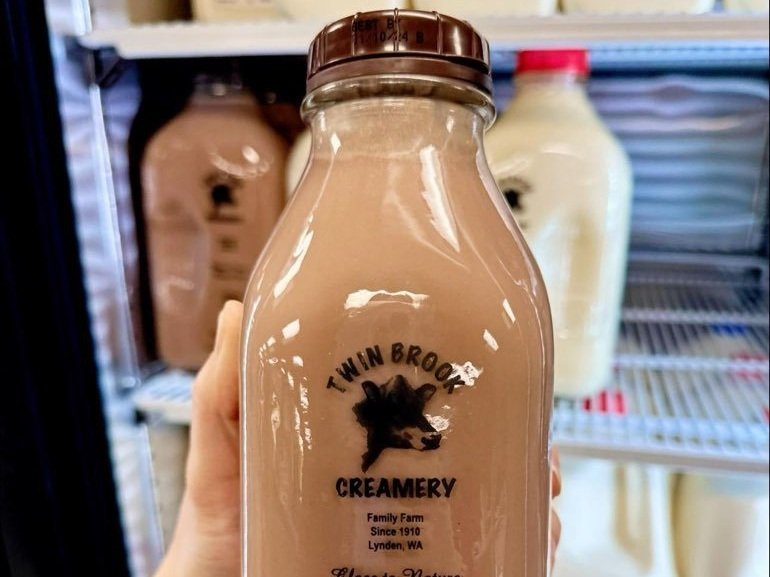 Brown glass bottle of chocolate milk from Twin Brook Creamery, a family farm established in 1910, displayed in a refrigerator. The label features a cow logo and indicates the product's origin in Lynden, WA.