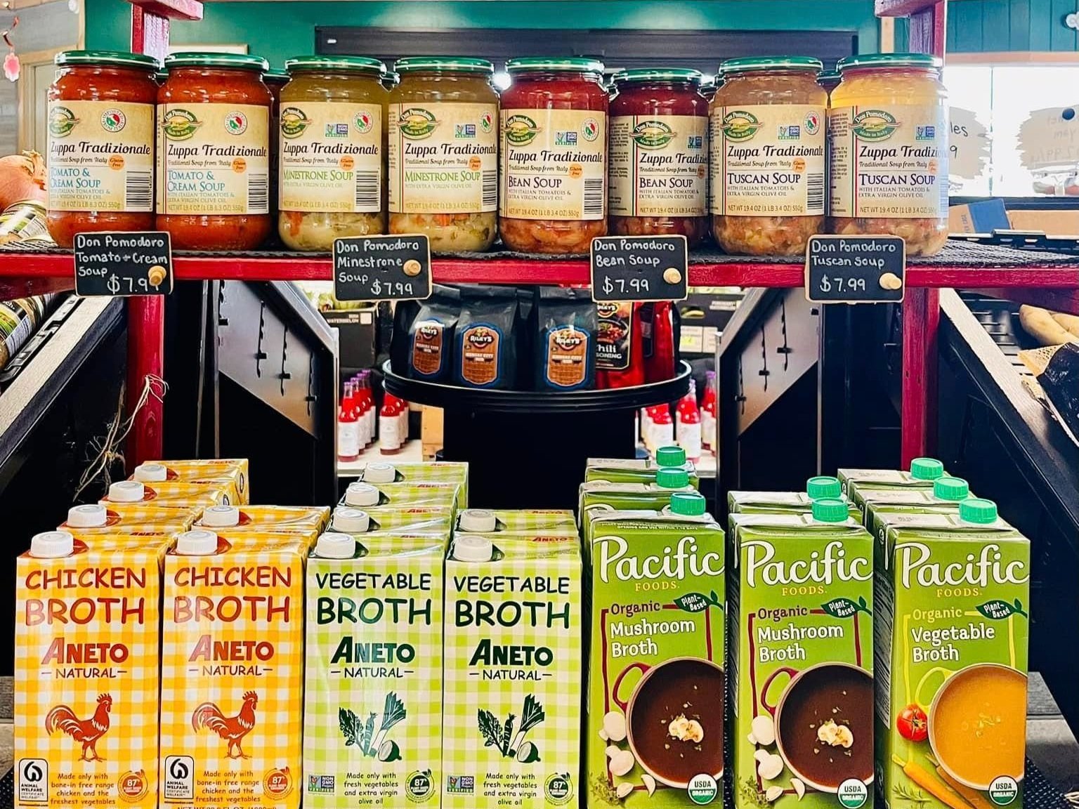 Shelves stocked with a variety of soups and broths, including Don Pomodoro Tomato & Cream Soup, Minestrone Soup, Bean Soup, and Tuscan Soup, alongside Aneto Chicken and Vegetable Broth, and Pacific Organic Mushroom and Vegetable Broth, all available for purchase.
