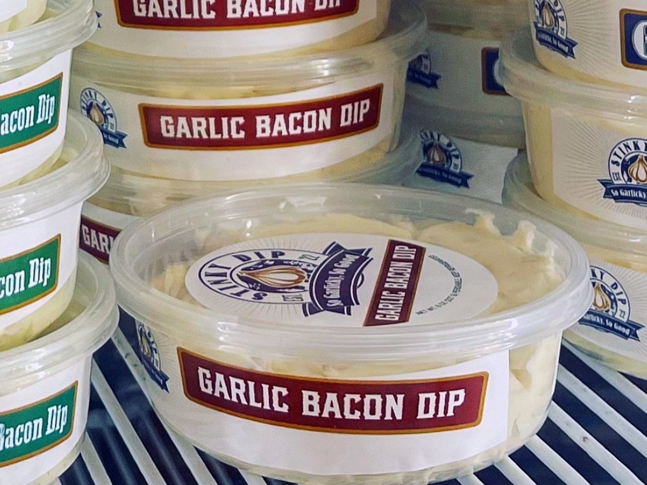 Containers of garlic bacon dip displayed in a refrigerated section, showcasing various labels and packaging styles. Ideal for appetizers or party snacks.