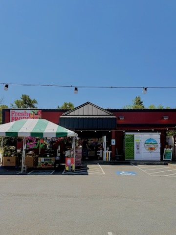 Exterior view of a fresh produce market featuring a colorful awning, vibrant signage, and a clear blue sky, with a parking lot in front.