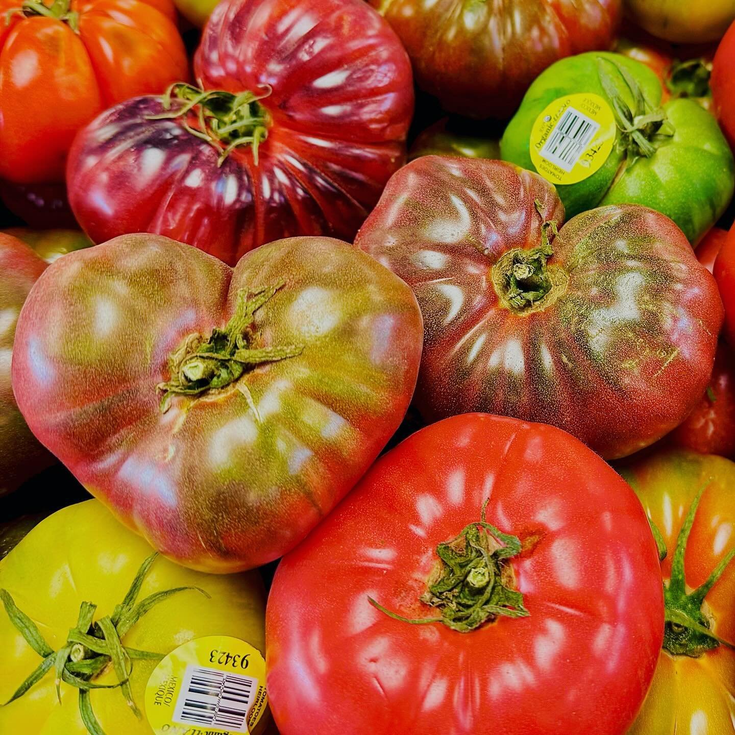 Patrick+Brown+-+Heirloom+Tomatoes (1) Colorful assortment of heirloom tomatoes, including heart-shaped and round varieties, showcasing vibrant red, green, and yellow hues, stacked together in a market display.