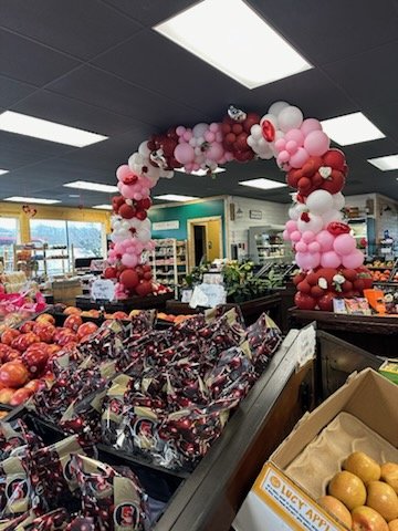 Colorful balloon arch decorated with pink and red balloons in a grocery store, creating a festive atmosphere for shoppers, with displays of fresh apples and packaged goods in the foreground.