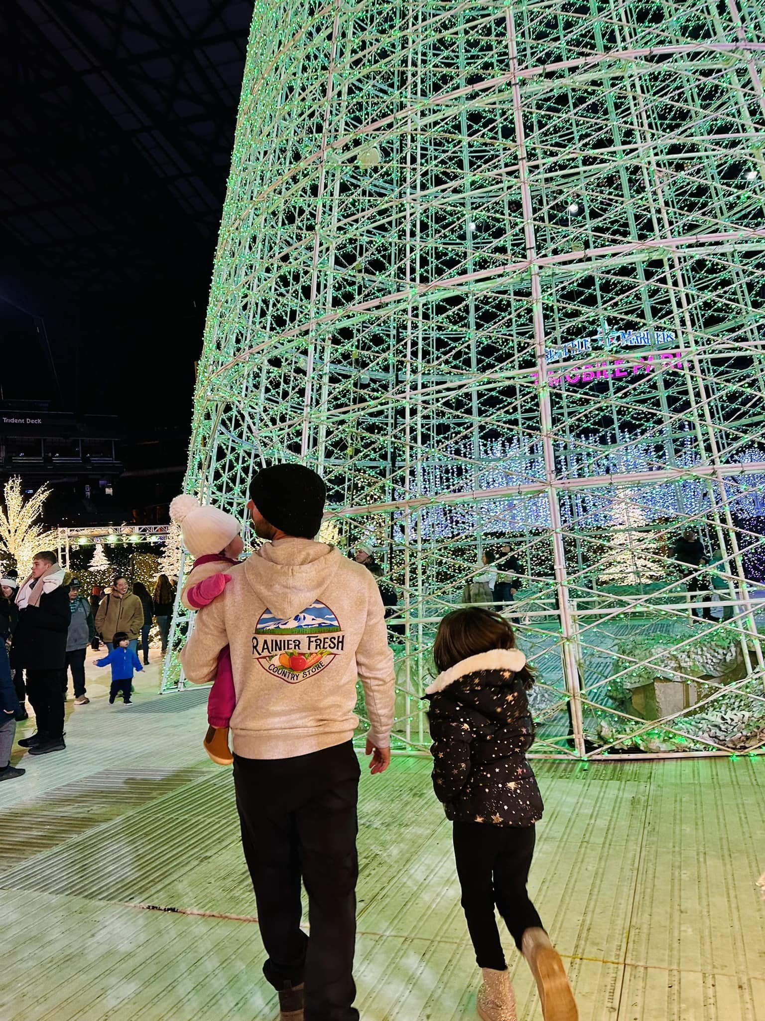 Family enjoying a festive holiday display at night, walking past a large, illuminated Christmas tree made of green lights, surrounded by other visitors in a winter wonderland setting.