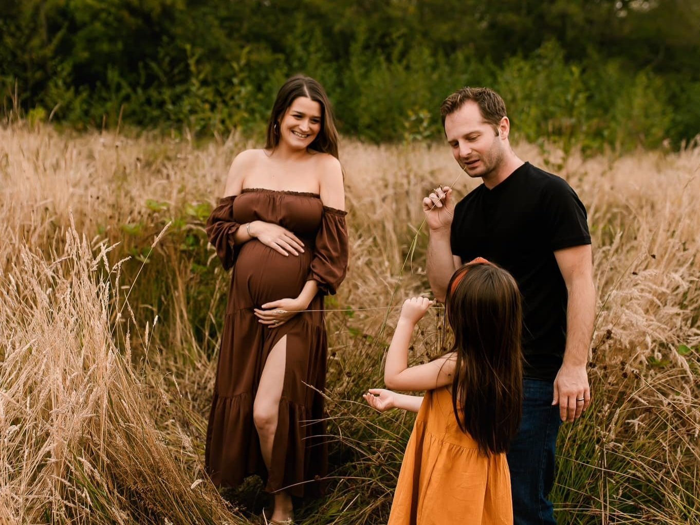 Pregnant woman in a brown dress smiling in a field, standing beside a man and a young girl. The family enjoys a moment together amidst tall grass and greenery, capturing the essence of love and anticipation for the new arrival.