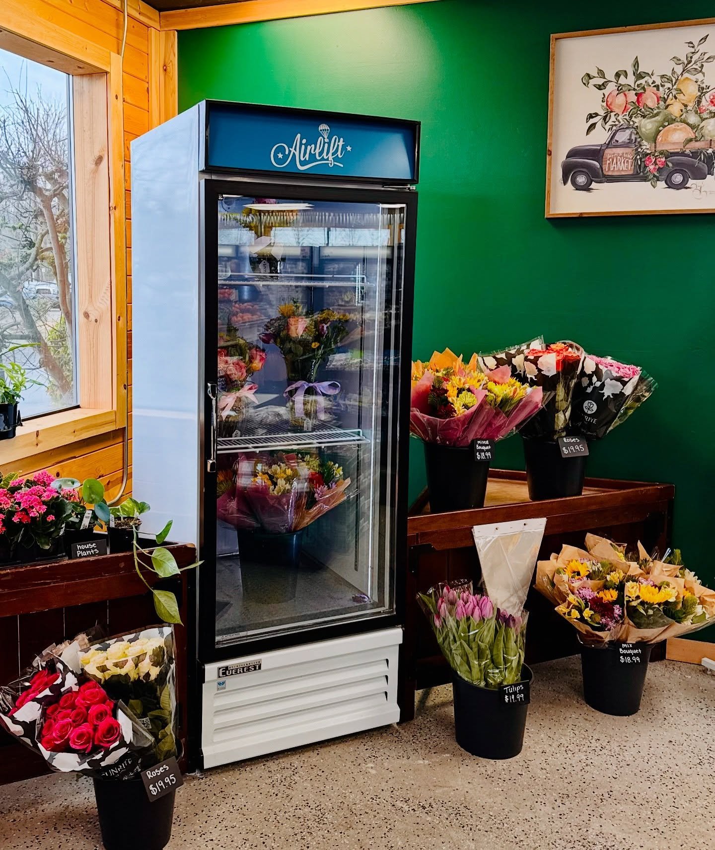 Refrigerated flower display showcasing a variety of colorful bouquets, including roses and tulips, in a floral shop with a green wall and wooden accents. The Airlift brand refrigerator highlights fresh flower arrangements for sale.