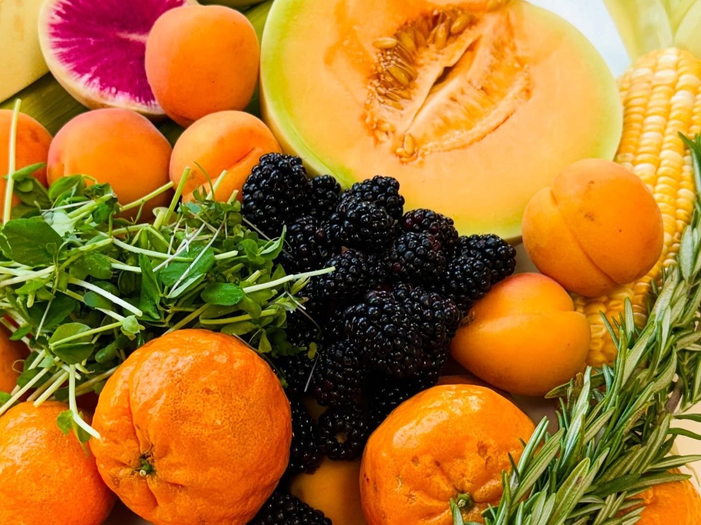 Colorful assortment of fresh fruits and vegetables including melons, apricots, blackberries, oranges, corn, and microgreens arranged on a platter, highlighting seasonal produce and healthy eating options.